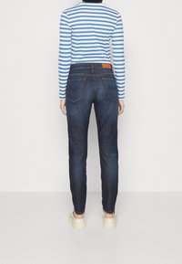  Jeans slim fit in denim scuro con due tasche posteriori, abbinati a una maglietta a maniche lunghe blu e bianca a righe. Le scarpe sono di colore chiaro e presentano un design arrotondato.