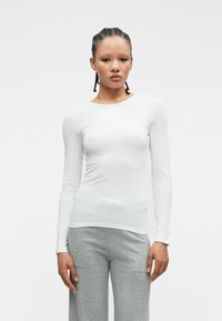 Biała, dopasowana bluzka z długim rękawem uszyta z gładkiego, elastycznego materiału. Okrągły dekolt, w połączeniu z luźnymi, jasnoszarymi spodniami. Minimalistyczny design.