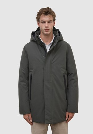 Young man wearing a dark green hooded winter coat with zippered pockets over a white shirt, standing against a plain light background.