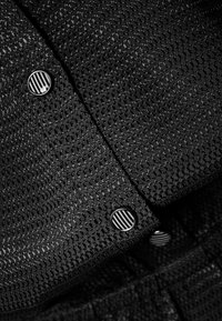 Tela negra texturizada con un patrón de malla hexagonal, que presenta botones redondos negros con un diseño de rayas blancas en la abertura.