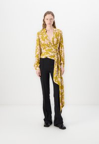 Gelbe Blumenkreppbluse mit langen Ärmeln, tiefem V-Ausschnitt und asymmetrischem Saum, kombiniert mit schwarzen, figurbetonten Hosen. Glatte Textur.