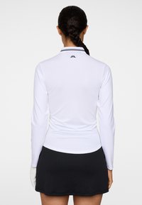 Polo à manches longues blanc avec une texture côtelée et un col noir orné d'un logo au dos, associé à une jupe plissée noire.