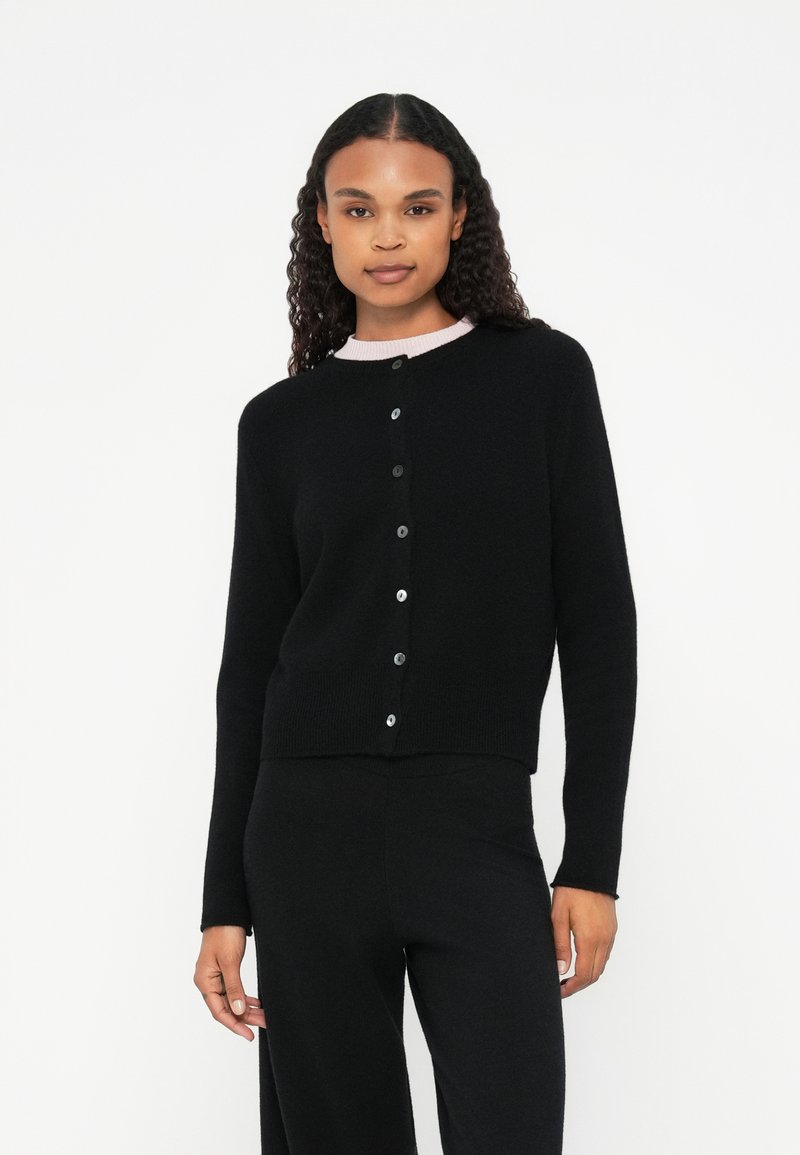 Cardigan preto com decote redondo, apresentando um acabamento em rosa contrastante. Tem um corte ajustado e seis botões na frente.