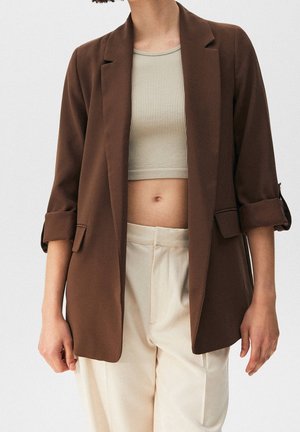 Hnědý blazer s přizpůsobeným střihem, dlouhými rukávy vyhrnutými nahoru a s klopami. Nosí se přes světlezelený ribbed crop top a béžové kalhoty.
