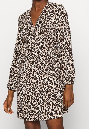 Kobieta w sukience kopertowej z długim rękawem i dekoltem w serek, z beżowo-czarnym nadrukiem w cętki i wiązaniem z boku w talii.