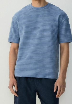 Uomo che indossa una maglietta a maniche corte di colore azzurro chiaro con texture e pantaloni blu scuro, in piedi con le mani rilassate lungo i fianchi.