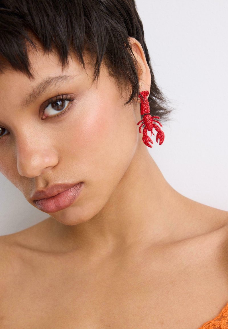 Jeune femme aux cheveux courts portant une boucle d'oreille rouge vif en forme de homard sur son oreille gauche, sur un fond clair uni.