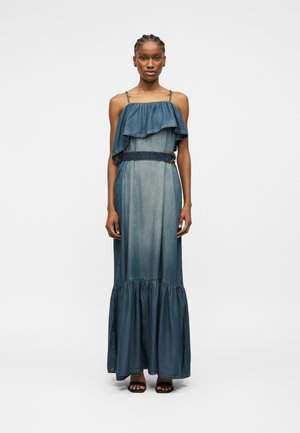 Žena v modrých rifľových maxi šatách bez rukávov s vrstveným volánovým topom a opaskom v páse, stojaca vzpriamene na obyčajnom pozadí.