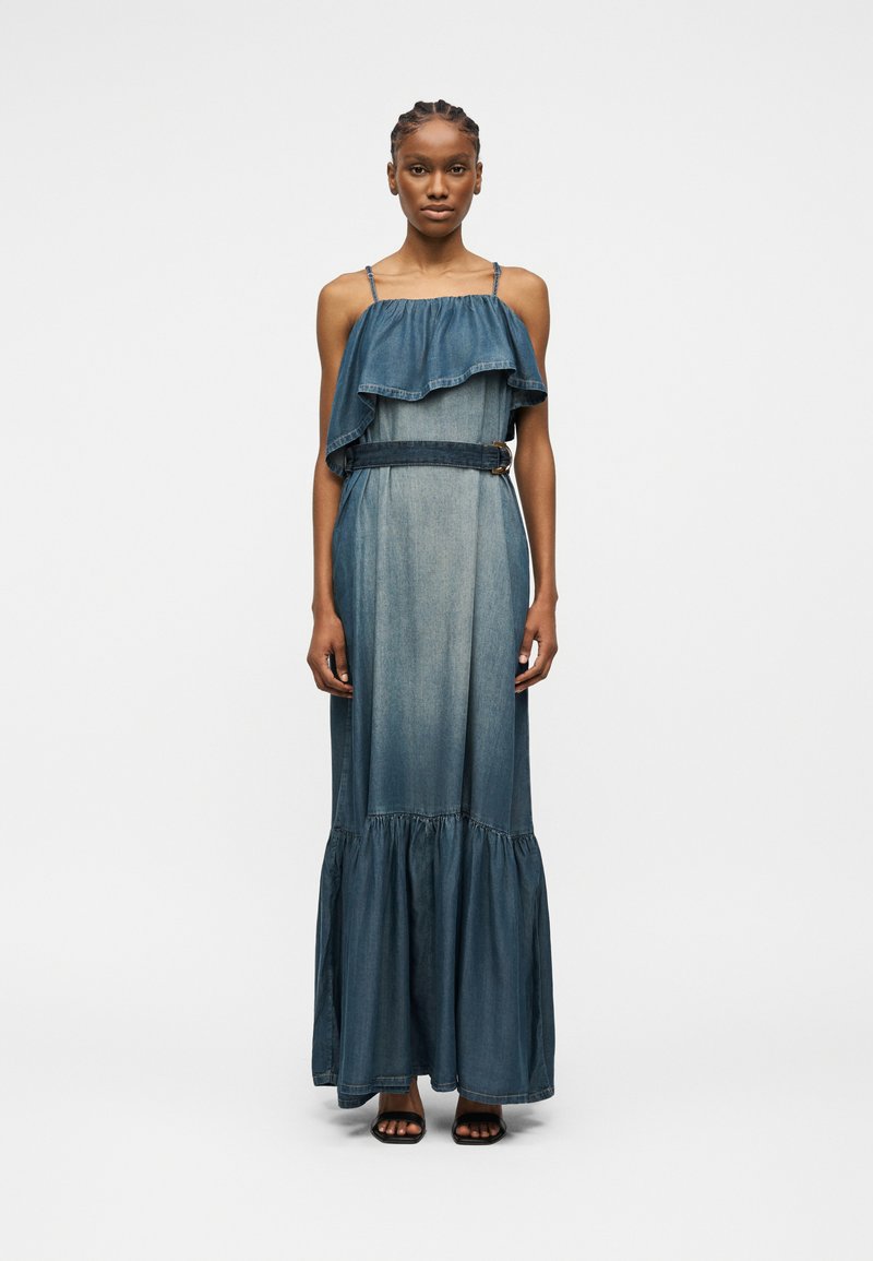 Γυναίκα που φοράει αμάνικο μπλε τζιν μακρύ φόρεμα με στρωμένες βολάν στο πάνω μέρος και ζώνη στη μέση, στέκεται όρθια μπροστά σε απλό φόντο.