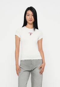 T-shirt en coton ribbed blanc à manches courtes, comportant un texte "tommyjeans" rouge et une broderie de rose sur la poitrine. Coupe classique ajustée.