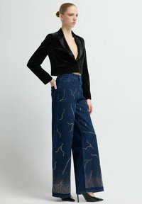 Blazer corto de terciopelo negro con adornos brillantes, combinado con vaqueros de mezclilla oscuros de pierna ancha que presentan patrones texturizados y detalles plateados.