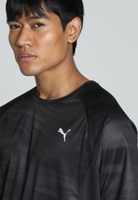 Sort atletisk T-shirt med korte ærmer, prydet med et tekstureret mønster og et hvidt Puma-logo på venstre bryst. Glat stof.