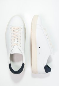 White leather sneakers with a smooth surface, round toes, cream laces, and a navy accent at the heel; textured rubber sole with small spikes.