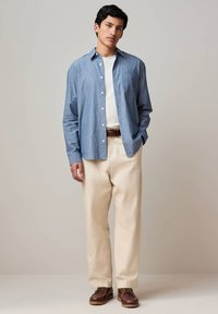Giovane uomo in piedi che indossa una camicia a righe blu con bottoni sopra una maglietta bianca, pantaloni beige, cintura marrone e scarpe di pelle marrone.