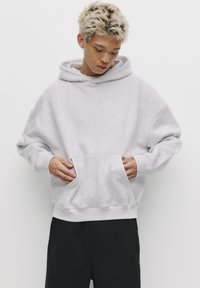 Jasnoszara, oversize bluza z kapturem, wykonana z miękkiego materiału. Posiada dużą kieszeń z przodu, ściągacze na rękawach oraz kapturem ze sznurkiem. Noszona z czarnymi spodniami.