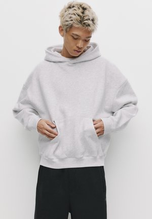 Hellgrauer übergroßer Kapuzenpullover aus weichem Stoff. Verfügt über eine große Fronttasche, gerippte Bündchen und eine Kordelzugkapuze. Kombiniert mit schwarzen Hosen.