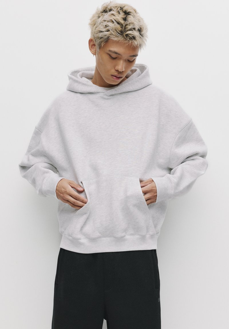 Jasnoszara, oversize bluza z kapturem, wykonana z miękkiego materiału. Posiada dużą kieszeń z przodu, ściągacze na rękawach oraz kapturem ze sznurkiem. Noszona z czarnymi spodniami.
