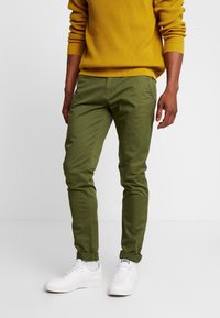 Gröna slim-fit chinos i bomull, med en mjuk yta, uppvikta cuffs och matchade med vita sneakers på en enkel bakgrund.