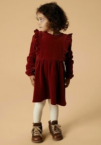 Rotes Samtkleid mit gerafften Schulterakzenten, langen Ärmeln und einem gerafften Bund. Kombiniert mit weißen gerippten Strumpfhosen und braunen Schnürstiefeln.