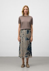 Vrouw met kort blond haar, gekleed in een taupe top met korte mouwen, een geplooide beige rok met donkerblauwe patronen en beige instapschoenen, die stil staat.