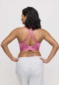 Reggiseno sportivo fucsia con spalline a forma di X, tessuto strutturato e chiusura a gancio. Pantaloni della tuta grigi visibili in vita. Vista posteriore.