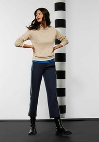 Beiger Pullover mit Rundhalsausschnitt und langen Ärmeln, kombiniert mit navyblauen verkürzten Hosen mit weißen Seitenstreifen. Schwarze Stiefeletten runden den Look ab.