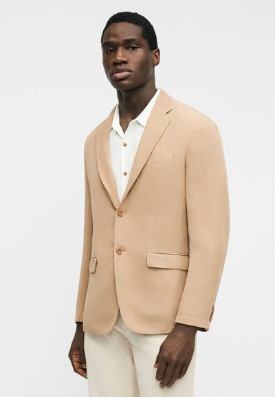 Άνδρας που φοράει μπεζ σακάκι πάνω από λευκό πουκάμισο με κουμπιά και ανοιχτόχρωμο παντελόνι, στέκεται μπροστά σε απλό ανοιχτόχρωμο φόντο.