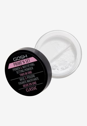 Récipient noir rond étiqueté "GOSH PRIME'N SET," contenant de la poudre translucide. Le couvercle présente des accents roses et un texte blanc. La base transparente montre le contenu.