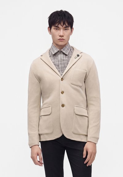 Млад мъж, облечен в бежово сако с три копчета върху карирана риза и черен панталон, стои пред бял фон.