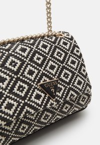 Black and white woven clutch with geometric pattern, gold chain strap, and a triangular Guess USA logo plaque on the front.
