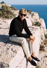 Homme blond portant des lunettes de soleil, une veste noire, un pantalon blanc et des mocassins noirs assis sur des falaises rocheuses avec la mer en arrière-plan.
