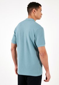 Gaiši zils polo krekls, īsas piedurknes, izgatavots no teksturētas auduma ar apmali un taisnu apakšu, skatīts no aizmugures.