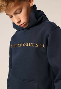 Marineblauwe hoodie met gouden tekst "DELUXE ORIGINAL," voorzien van een buidelzak en een zachte textuur.