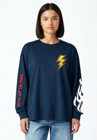 Femme portant un t-shirt manches longues bleu marine avec un éclair jaune sur la poitrine, "Vous avez le pouvoir" en rouge sur la manche gauche, et des flammes blanches sur la manche droite.