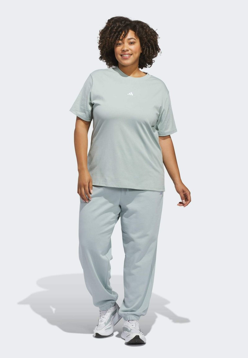 Mujer sonriente de pie, vistiendo una camiseta Adidas gris claro, pantalones deportivos a juego y zapatillas blancas sobre un fondo blanco.