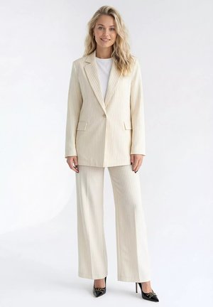 Blondynka ubrana w kremowy garnitur w cienkie prążki z szerokimi spodniami i marynarką na jeden guzik, w połączeniu z czarnymi szpiczastymi szpilkami, stoi i się uśmiecha.