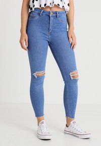 Jeans pitillo de talle alto en azul con rodilleras desgastadas, que presentan un diseño clásico de 5 bolsillos y un dobladillo desflecado, combinados con zapatillas blancas.