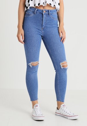 Jeans pitillo de talle alto en azul con rodilleras desgastadas, que presentan un diseño clásico de 5 bolsillos y un dobladillo desflecado, combinados con zapatillas blancas.