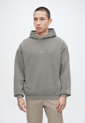 Lühikeste juustega mees, kes kannab halli kapuutsiga dressipluusi ja beeže pükse, seisab ühevärvilise valge tausta ees.