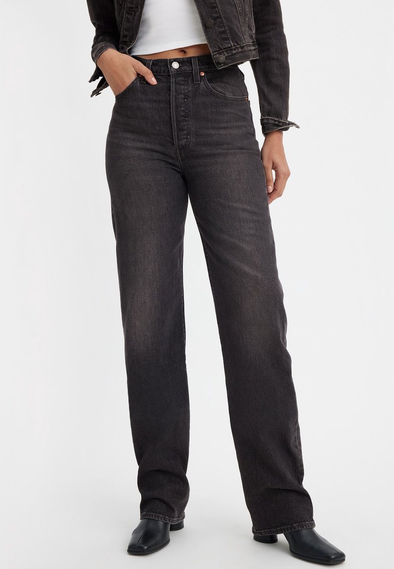 Femme portant un jean noir taille haute à jambes droites, un top blanc court, une veste en jean noire courte et des bottines noires, debout avec une main dans la poche.