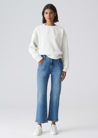 Felpa bianca in pile con maniche ampie, abbinata a jeans flare azzurri chiari con tasche frontali e orlo grezzo, indossata con sneaker bianche.