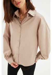 Chemise beige clair à boutons avec un col à volants et de longues manches bouffantes ; le tissu semble lisse avec un éclat subtil.