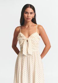 Kremowa sukienka z dopasowanym korpusem, zdobieniami w postaci marszczenia, kokardą z przodu oraz zwiewną spódnicą, z czarnymi kropkami na całej powierzchni.
