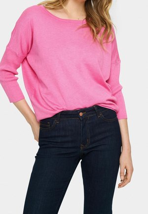 Femme portant un pull rose à manches longues ample rentré dans un jean taille haute bleu foncé, main posée sur la hanche.