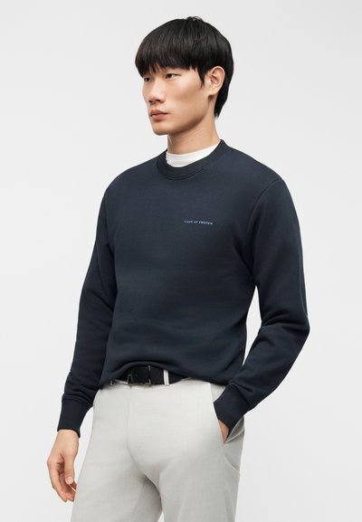 Μπλε ναυτικό φούτερ με στρογγυλή λαιμόκοψη, με μικρό ανοιχτό μπλε λογότυπο "Tiger of Sweden" στο στήθος; φορεμένο με ανοιχτό γκρι παντελόνι.