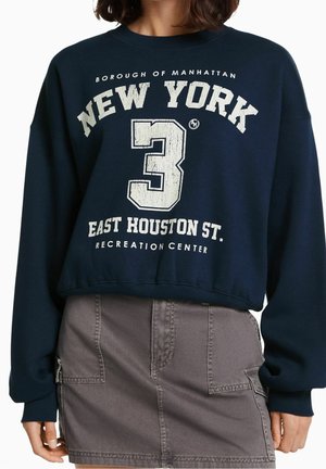 Sweat-shirt bleu marine avec texte blanc usé "New York 3 East Houston St. Recreation Center", porté avec une jupe grise munie de poches frontales.