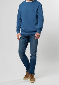 Felpa blu a collo rotondo con polsini e orlo a costine, abbinata a jeans blu attillati e sneakers color beige. Piccolo logo sul petto.