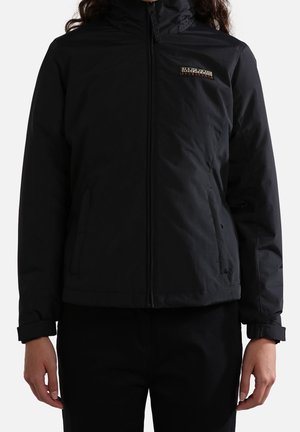 Osoba ubrana w czarną kurtkę na zamek z wysokim kołnierzem i małym prostokątnym logo na lewej piersi, stojąca z rozluźnionymi ramionami.