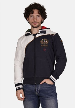 Giacca con zip navy e bianca con cappuccio; presenta ricami, polsini a righe e una combinazione di colori solidi su maniche e collo.