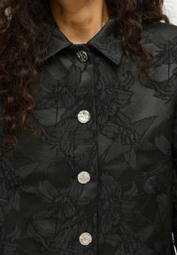 Veste à motif floral noir avec de grands boutons argentés, un col pointu et une texture lisse avec un éclat subtil.